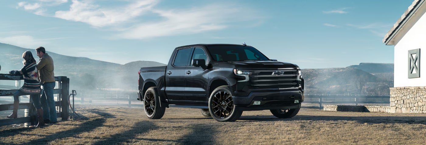 Image of a black 2025 Silverado 1500 Highcountry parkec on a rustic ranch while a rancher couple leans against a fence